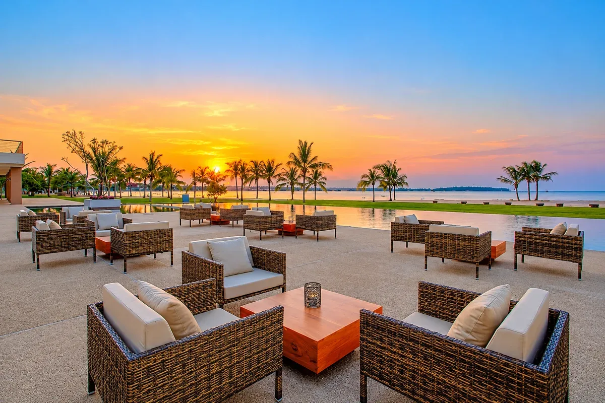 Outdoor seating beside a pool overlooking palm trees and the sea at sunset