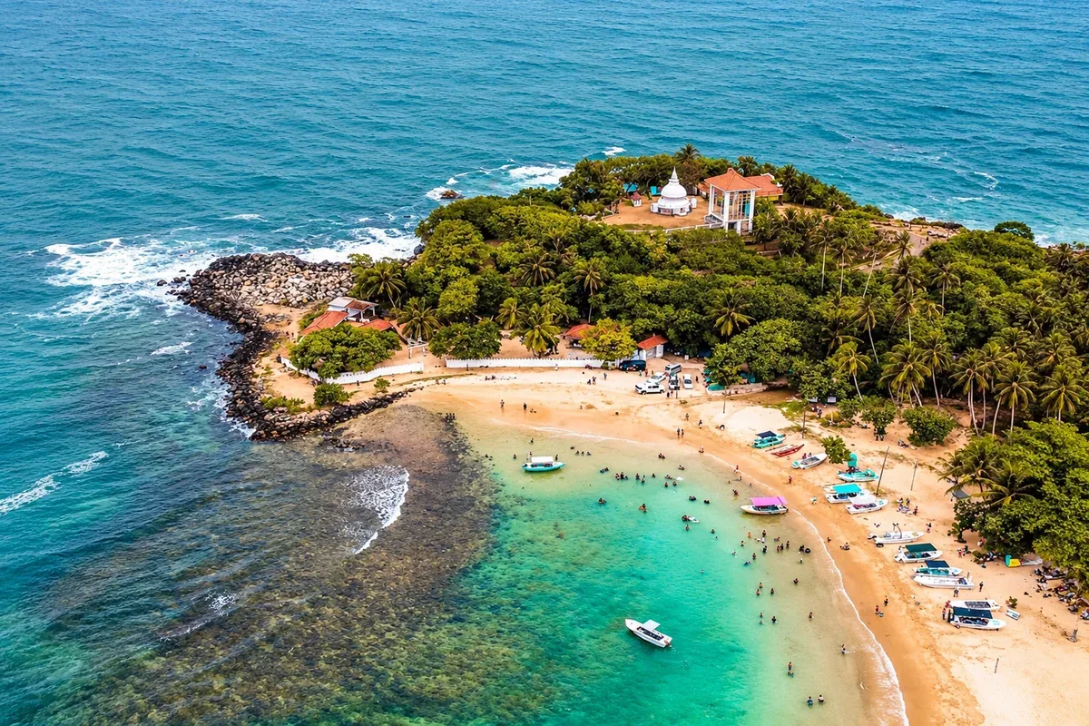 Aerial view of a sandy bay with boats, swimmers and a temple on a green headland